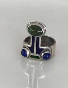 There is a hint of arts and crafts, as well as, modernist in this unusual ring which you can wear with the Peridot facing up or down! The beautiful green stone, as well as the smaller vibrant blue Lapis are all cabochon and bezel set. The enamel is in wonderful condition. Aside from some slight wear, the ring is in overall fabulous shape. The band is almost 1/4" wide, it is marked Sterling and has the initials CHM scratched on the silver behind the Peridot. Please see all photos for details, as Lapis And Peridot, Beautiful Rings Vintage, Old Rings, Unusual Rings, Geometric Ring, Peridot Ring, Peridot Gemstone, Gold Band Ring, Blue Lapis