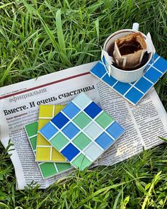 a newspaper laying in the grass next to a cup of coffee and some colorful tiles