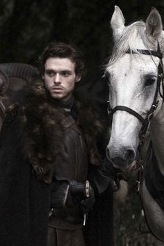 a man standing next to a white horse wearing a black coat and fur stole around his neck