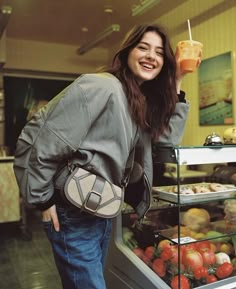 a woman holding a drink in her right hand and standing next to a display case full of food