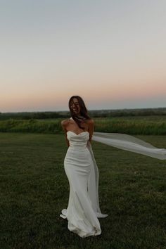 a woman in a white dress is standing on the grass with her veil blowing in the wind