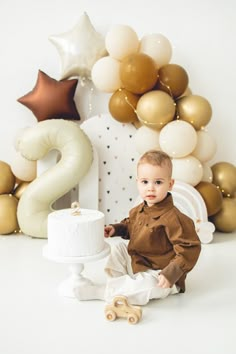 a baby boy sitting on the floor next to a white cake with gold balloons in the background