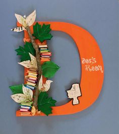 an orange letter made out of books with leaves and butterflies on it, sitting in front of a blue wall