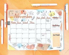 an open planner with flowers and leaves on it, next to two pens that are resting on a wooden surface