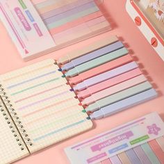 various stationery items are laid out on a pink surface, including notebooks and pencils