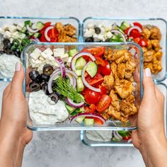 two hands holding up a glass container filled with salad and chicken, cucumbers, tomatoes, onions, olives, lettuce