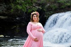 a pregnant woman standing in front of a waterfall wearing a pink dress and flower crown