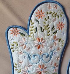 two embroidered oven mitts are hanging on a white cloth covered wall with blue trimmings