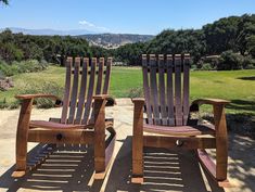 two wooden lawn chairs sitting on top of a stone patio next to a lush green field