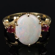 Pretty raspberry red rubies flank a natural white 1.68 carat Australian opal on this vintage 14k yellow gold ring. At the center is an oval 12mm x 9.8mm x 2.7 opal cabochon of white body and an array of color flashes. Fiery sparkles of greens, blues, reds and yellows come through when the gem refracts the light. The camera did not quite capture the actual beauty of the stone. The faceted 3mm pinkish-red rubies have an estimated total weight of .20 carat. The opal is held tightly with six sturdy Large Opal Ring, Oval Ruby Ring, Pinkish Red, Raspberry Red, Ruby Jewelry, Red Ruby, Australian Opal, Yellow Gold Ring, Ruby Ring