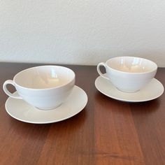 two white cups and saucers sitting on top of a wooden table next to each other