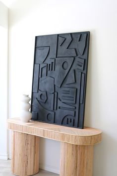 a wooden table topped with a vase next to a black art piece on top of it