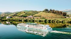 a large white boat traveling down a river next to a lush green hill covered hillside