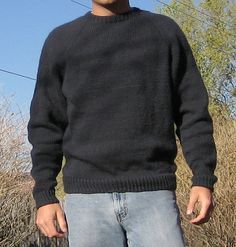 a man standing in front of a fence wearing a sweater and jeans with his hands in his pockets