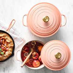 two pink pots with food in them on a white counter top next to a pot filled with fruit