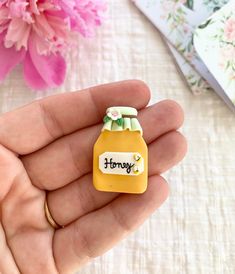 a hand holding a tiny honey jar with the word honey written on it and flowers in the background