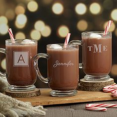 three mugs with hot chocolate and candy canes on a wooden tray in front of christmas lights