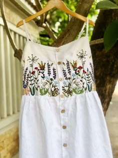 a white dress hanging on a tree with flowers and plants embroidered on the chestline