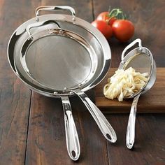 two stainless steel pans on a wooden cutting board with tomatoes and garlic in the background