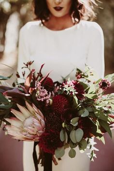 a woman holding a bouquet of flowers and greenery in front of her face, wearing a white dress