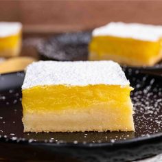 two slices of lemon bars on a black plate with sugar sprinkled around them