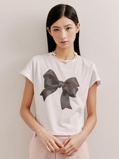 This is a short-sleeved, crewneck t-shirt that features a large bow graphic on the front. The graphic gives the impression of a textile bow tied at the center, adding a decorative and feminine touch to the classic tee. The shirt's design is simple and clean, making it versatile for various occasions. The combination of the casual cut and the eye-catching graphic creates a playful yet chic garment.- The bow graphic serves as a bold statement piece on the otherwise minimalist t-shirt.- Its classic crewneck and short-sleeve design make it a staple piece for any casual wardrobe.- The contrast between the graphic and the shirt suggests a design that's both modern and stylish. Trendy Summer T-shirt With Bow, Trendy Crew Neck T-shirt With Bow, Casual Crew Neck T-shirt With Bow Print, Spring Cotton T-shirt With Bow, White Short Sleeve Tops With Bow, Chic Spring Tops With Ribbon, Spring Crew Neck T-shirt With Bow, Cotton Summer Tops With Bow Print, Trendy White Top With Bow