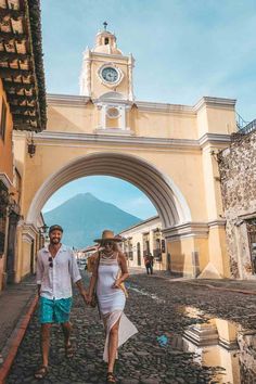 a man and woman walking down the street in front of an arch with a clock on it