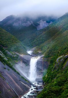 the waterfall is surrounded by lush green trees and foggy mountains in the distance, with low hanging clouds