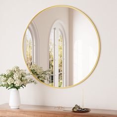 a white vase with flowers sitting on top of a wooden table next to a round mirror