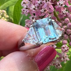 a woman's hand holding an aquamarinee and diamond ring in front of purple flowers