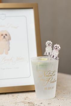 two small white dogs sitting on top of a glass next to a framed photo and candle