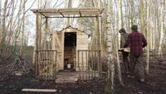 a man standing next to a small wooden structure in the woods