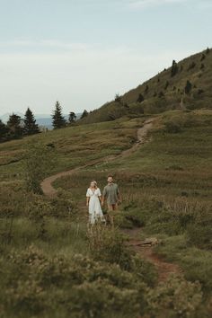 a man and woman walking down a dirt path in the grass on top of a hill