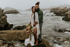 a man and woman standing on rocks near the ocean with their arms around each other