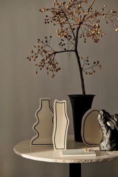 a white table topped with black and white vases next to a small tree filled with leaves