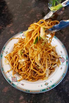 a plate full of noodles and vegetables with tongs sticking out of the top, ready to be eaten