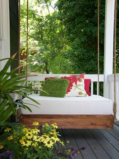 a porch swing with pillows on it in front of some flowers and trees, along with potted plants