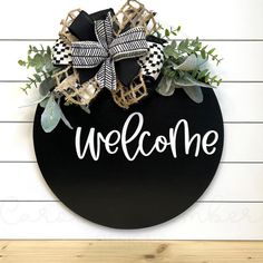 a welcome sign hanging on the side of a white wall with greenery and burlocks