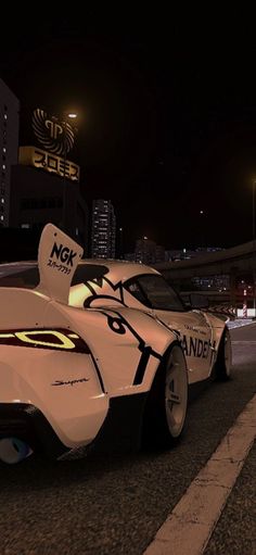 a white race car driving down a city street at night with lights on the buildings in the background