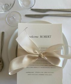 a table set with silverware, wine glasses and a white dinner plate that has a ribbon on it