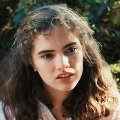 a close up of a person wearing a white shirt and long hair with trees in the background