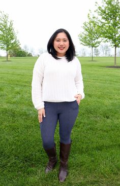 a woman standing in the grass with her hands in her pockets and smiling at the camera