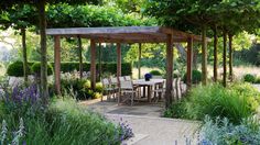 a table and chairs under a pergoline surrounded by trees in the middle of a garden