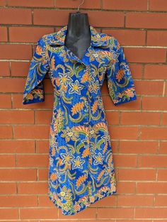 Found! 🔍 Super cool shirt-dress with asymmetrical button line and ability to be buttoned high necked or folded open. We especially love the cobalt blue colour contrasted with the orange tropical paisley floral swirling all over. 📋 - This dress is in excellent vintage condition with no noticeable defects. Made by M3rlin.  Size S.  👗 Measurements (taken flat):  - 93cm length  - 40cm shoulder  - 44cm bust  - 38.5cm waist  - 35.5cm sleeve length  - 15cm cuff width ------------------------------ Blue Short Sleeve Dresses With Buttons, Blue Button-up Shirt Dress For Work, Blue Knee-length Dress With Button Closure, Blue Button-up Shirt Dress For Daywear, Blue Stand Collar Summer Dress, Blue Collared Shirt Dress With Placket, Casual Blue Shirt Dress With Placket, Blue Summer Dress With Stand Collar, Blue Dress With Stand Collar