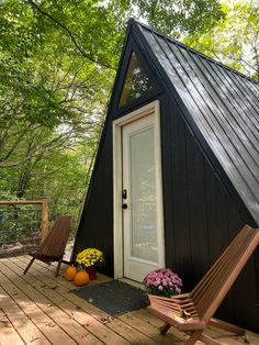 a small black cabin in the woods with two chairs and flowers on the front porch