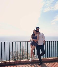 a man and woman standing next to each other on a balcony near the ocean with their arms around each other