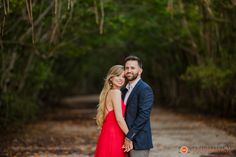 a man and woman standing in the middle of a dirt road with trees behind them