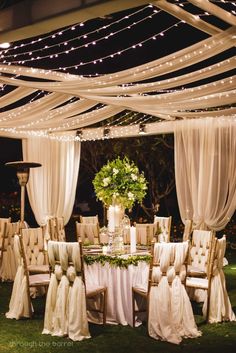 an outdoor wedding setup with white draping and lights on the ceiling, decorated with flowers and greenery