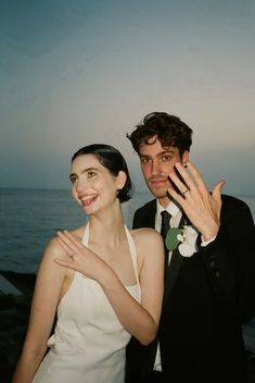 a man and woman standing next to each other in front of the ocean with their hands up