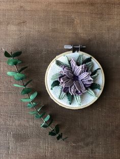 a purple flower is on the table next to some green leafy branches and a small cross - stitch pattern